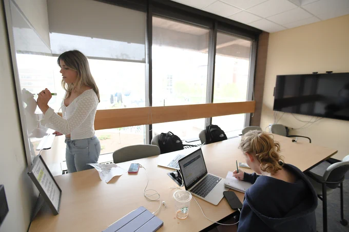 Two students are studying in a private study room. One student is writing on a white board at the front of the room and another is sitting at a table and taking notes.