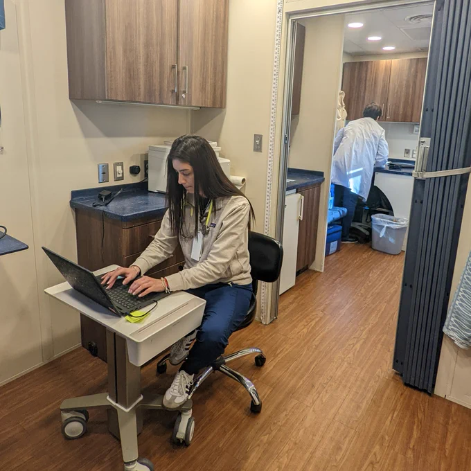 Community Care Coach team member sits at a desk inside the Care Coach and works on a laptop. Directly behind them is another another team member with their back facing the camera from another room.