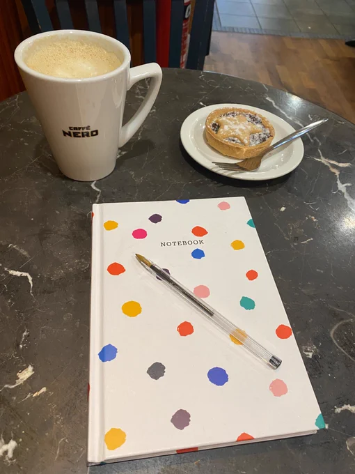 Photo shows a table in the coffee shop Caffè Nero. On the table is a large mug of coffee, a mince pie on a plate, and a white notebook with colourful polka dots. 