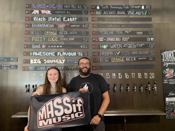 Two smiling members of the Torchlight team hold a Massif banner in front of a big board listing beer and drink options