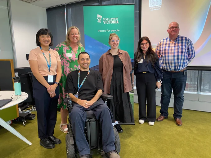 Six people in a group with the Development Victoria banner located behind them. One person is seated in a power wheelchair and the others are standing. All are smiling at the camera.