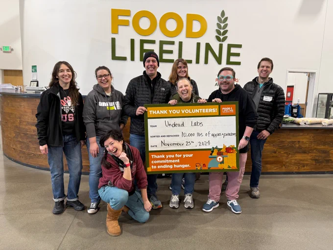 Group of people smiling and holding a large check in front of a "Food Lifeline" sign. The check, from Wheel Labs, donates 100,000 lbs of paper products.