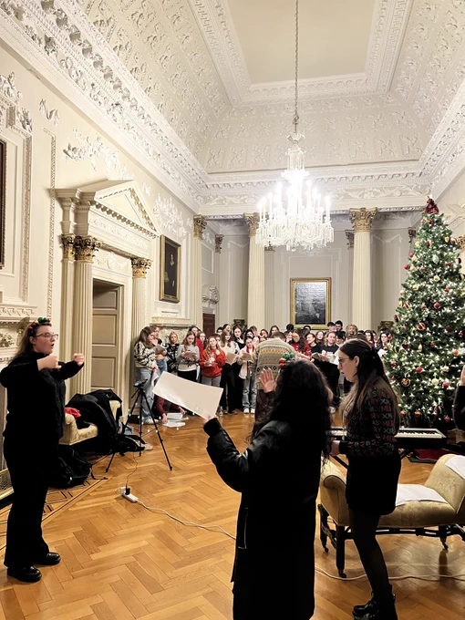 Wide shot taken in a big room. There is a choir singing beside a tall Christmas tree in the background, and ISL interpreters signing in the foreground.
