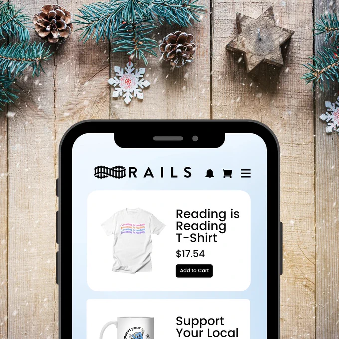 A wooden background with pine, acorns, and snowflakes surrounding it. A phone with its screen displaying a Rails store with a t-shirt that says “reading is reading is reading” and a mug that reads “support your local library.” 