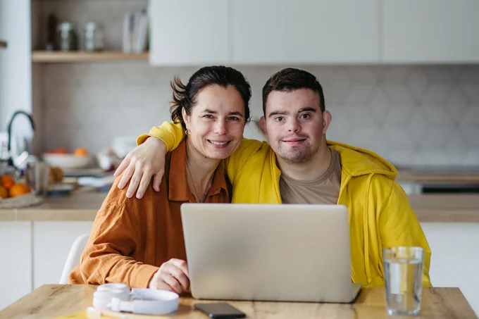 Mother with her son with disability in home kitchen, looking at a laptop.