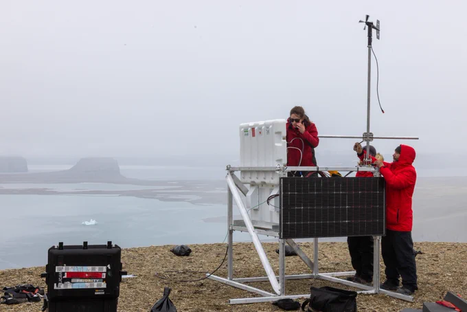 Trois personnes revêtant des manteaux d’hiver travaillent sur une structure munie d’un cadre en métal d’environ deux mètres cubiques et d’un panneau solaire sur la paroi. Des fils relient un couvercle ouvert à la boîte principale sur la structure. Une petite hélice se trouve sur le dessus d’un poteau fixé sur la structure.