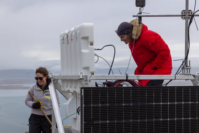 Une personne revêtant un manteau d’hiver travaille sur une structure munie d’un cadre en métal et d’un panneau solaire sur la paroi. Des fils relient un couvercle ouvert à la boîte principale sur la structure. Un poteau est fixé sur la structure. Une deuxième personne tient le manche de bois d’un outil près de la structure.
