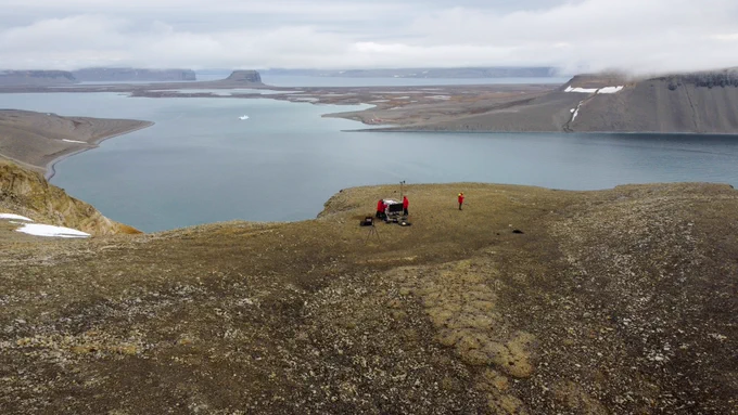 A drone shot from above of a structure about 2 cubic metres in size. Three people work on the structure. In the background, there is a body of water with high cliffs with some snow patches. A cluster of several buildings is near the water in the distant background.