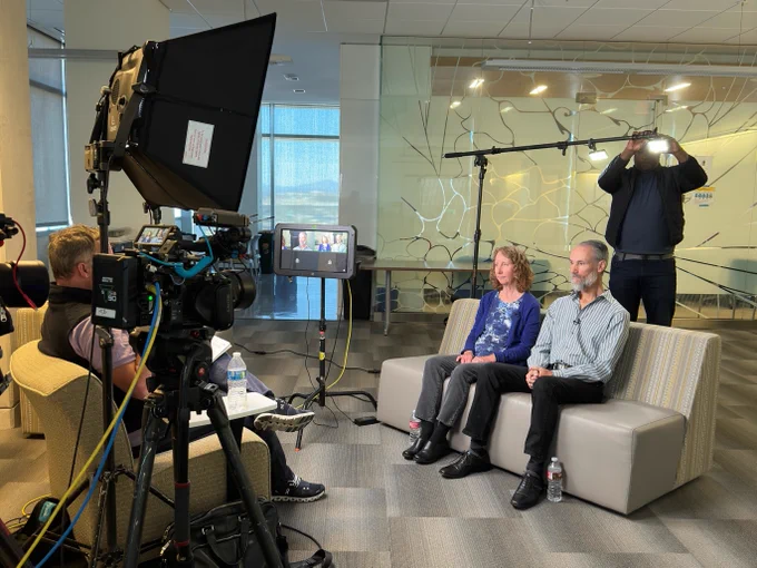 Peter Lucchini and his wife are seated on a sofa being interviewed in a professional studio setting with cameras, lights, and a production crew.