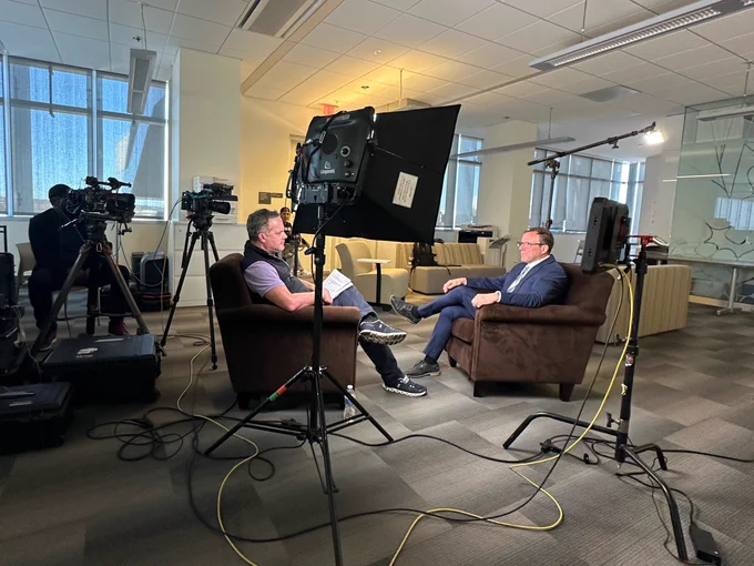 Dr. Gabriel Leger and an interviewer in a television studio setup, seated in armchairs facing each other, with cameras, lights, and a backdrop of a room with large windows.