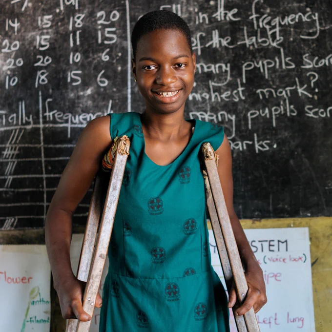 A schoolgirl with crutches smiles as she stands in her classroom.