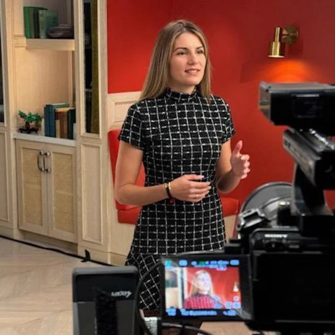 A person with shoulder length straight blond hair wearing a black and white checkered dress gestures while being filmed by a professional camera. The person is in a room with a red wall and a bookcase. A monitor shows the camera's view of the person
