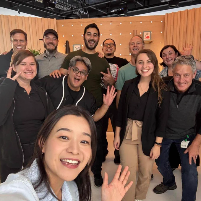 A group of people smile and wave for a selfie. They are standing in a room with light-colored walls and wooden accents. 
