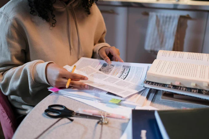A person sitting at a desk with an open textbook in front of them, while gazing at a few sheets of notes or articles.