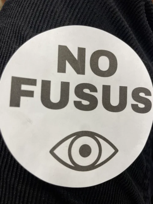 A white, circular sticker that reads “No FUSUS” with an image of an open eyeball.
