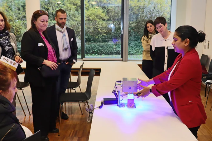 A woman in a red blazer explains a technical device on a table emitting blue light to a small group. The attendees watch the demonstration attentively in a modern room with large windows.