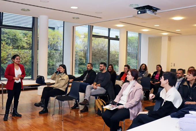 A woman in a red blazer is giving a presentation to a small group of attendees in a modern seminar room with large windows. The audience is seated on chairs and listening attentively.