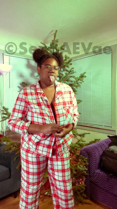 Vae is standing in her living room wearing red plaid, Christmas pjs sticking her tongue out at the camera.