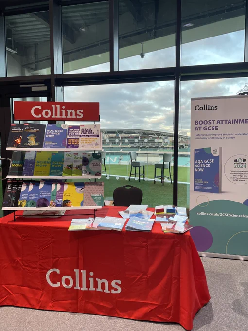 Image shows a table covered in a red Collins branded tablecloth, with a book stand displaying three shelves of Collins books. The background shows the Oval Cricket ground