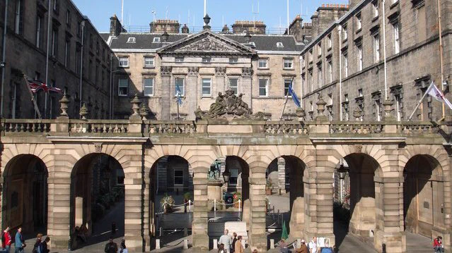 The facade of the Edinburgh City Chambers on a sunny day. People are walking past and flags are flying from the roof of the building. 