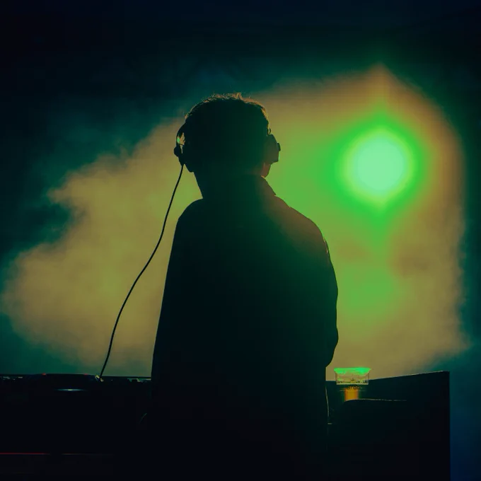 A silhouette of a DJ wearing headphones, standing at a deck with a bright green light and atmospheric smoke in the background.