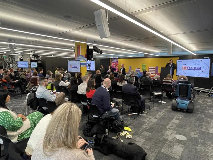 A wide shot of Gary O’Donoghue welcoming people to the Rethink Disability event. Gary is seen standing behind a podium, wearing a dark suit, with a screen showing the BBC Rethink Disability Festival logo shown, to his left. People are sitting in the audience in front of him and we can see the backs of their heads.