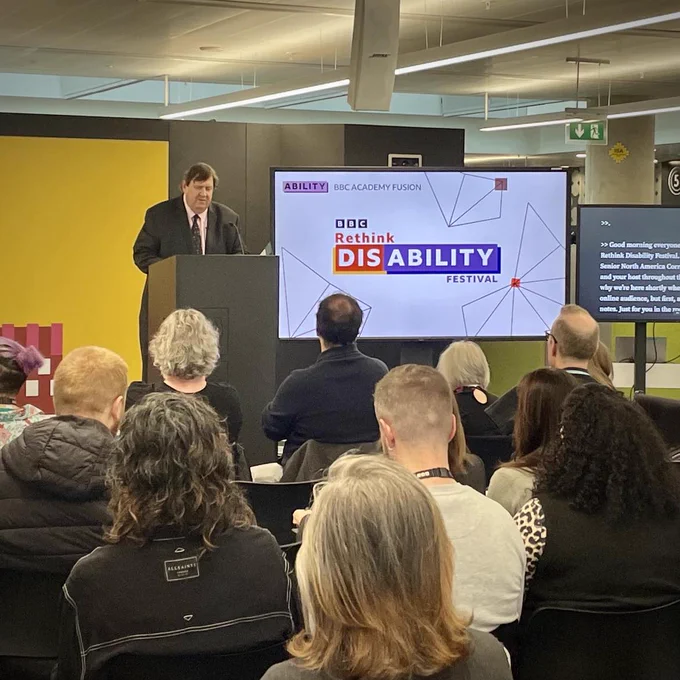 A photo of Gary O’Donoghue hosting the Rethink Disability event. Gary is seen standing behind a podium, wearing a dark suit, with a screen showing the BBC Rethink Disability Festival logo shown, to his left. A live caption screen is seen beyond that. People are sitting in the audience in front of him and we can see the backs of their heads.