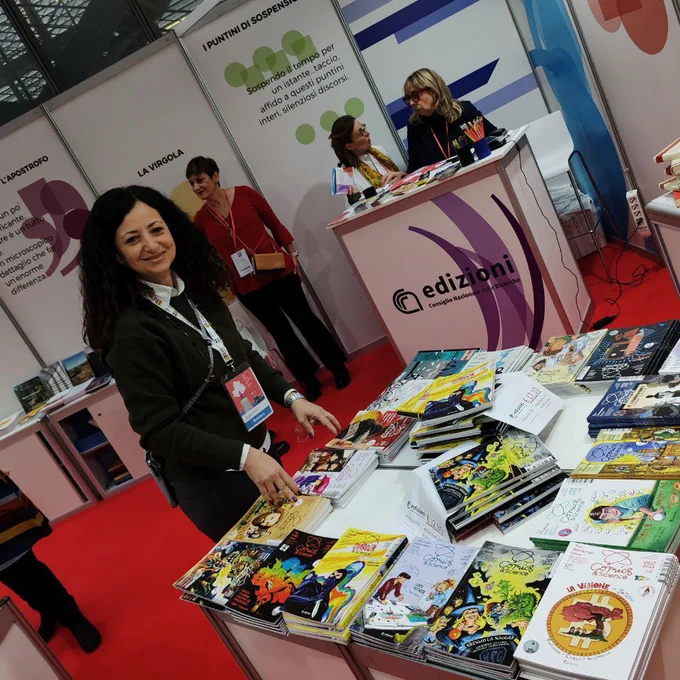 Una donna sistema degli albi a fumetti su un tavolo in uno stand fieristico. Si tratta di Stefania Giuffrida, CNR-IAC, e gli albi sono firmati Comics&Science, editi da CNR Edizioni. La foto è stata scattata all'interno della Nuvola a Roma, per la fiera della piccola e media editoria: Più Libri Più Liberi.