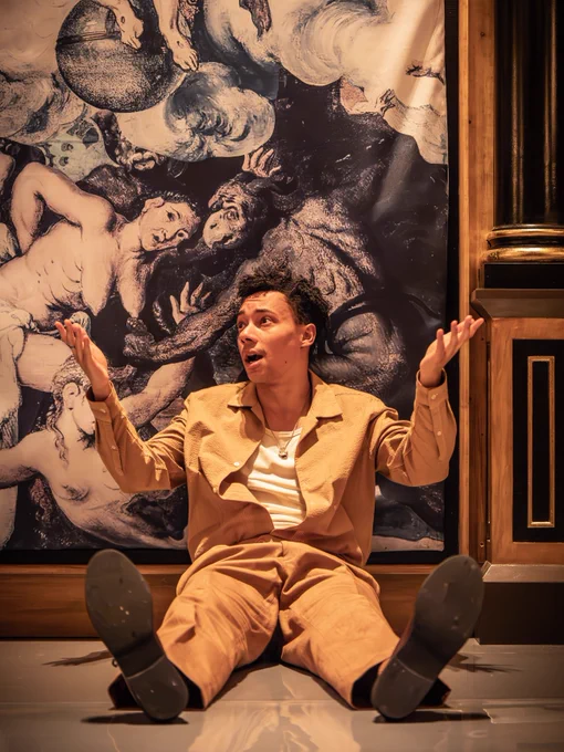 Kit Young as Bertram in All's Well that Ends Well (2024/5). 

An actor sitting on the floor, posing dramatically with an artistic mural of figures swirling in a blue background, in the background. The actor is dressed in a beige shirt, white shirt, and beige pants.
