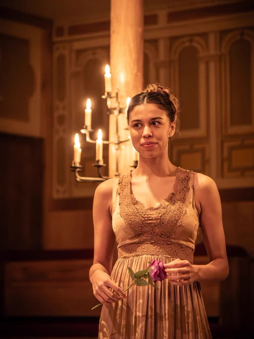 Georgia-Mae Myers as Diana in All's Well That Ends Well (2024/5).

An actor standing on stage in the Sam Wanamaker Playhouse. She is holding a flower, with candlelight and ornate architectural details in the background.