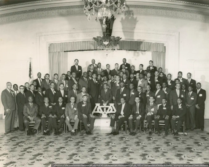 A black-and-white photograph of Alpha Phi Alpha fraternity members. The men are arranged in several rows on and in front of a curtained stage with a large Alpha Phi Alpha sign at front center. The uncropped shot shows several large chandeliers hanging from the ceiling and the rose-printed carpet of the room. On the verso are two stamps in blue ink, one reads [APR 21 1962] and the other reads [Burton's Studio / 12181/2 North Kenosho / Tulsa 6, Oklahoma / LUther 7-1463].
