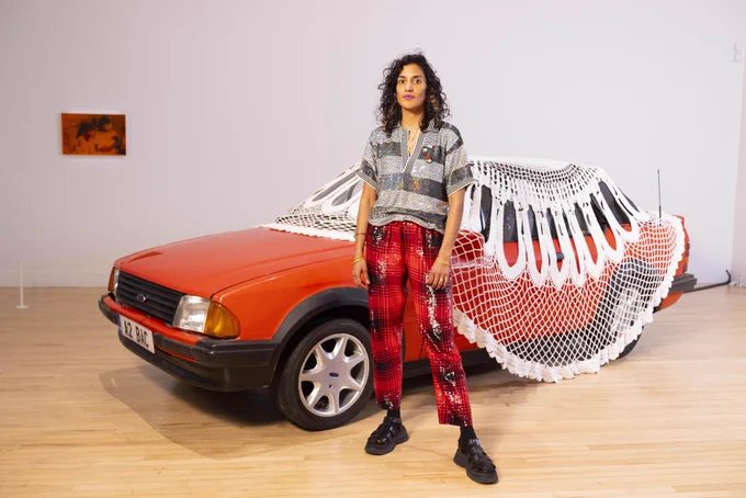 A woman wearing shiny plaid trousers and a sparkly striped top stands in front of a red Ford Escort with a car sized doily on top