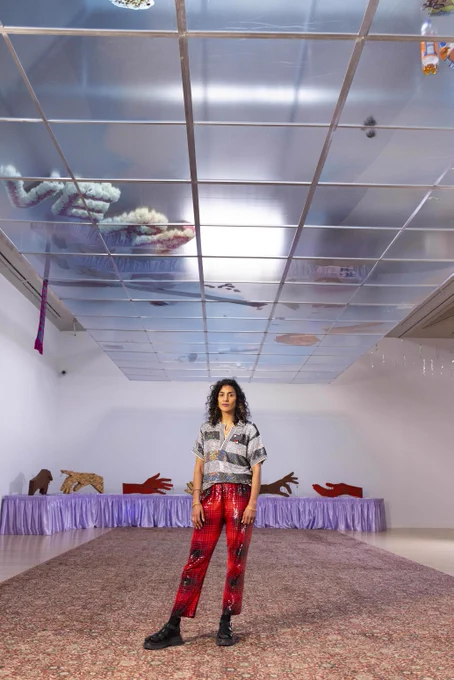 A woman wearing shiny plaid trousers and a sparkly striped top stands in a room with a translucent ceiling, there are sculptures of hands behind her