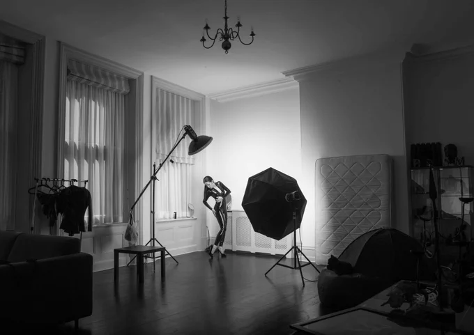 A black and white photograph of what looks like a large living room space. There is the corner of a sofa in the bottom left corner of the frame, an iron candle chandelier and a collection of trinkets in the right of the frame. In the centre of the room, a person wearing a dark jumpsuit poses in the spotlight created by two pieces of photography lighting equipment.