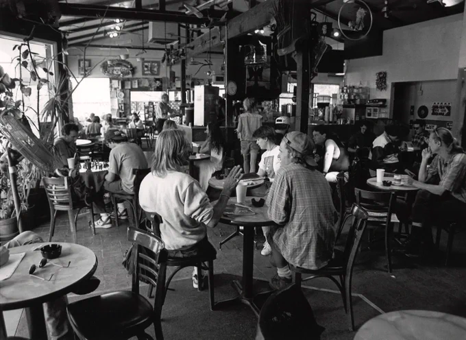 A black and white photo of the interior of Penny Lane Coffeehouse. Patrons sit at small circular tables. Most are engaged in conversation, two appear to be playing chess.