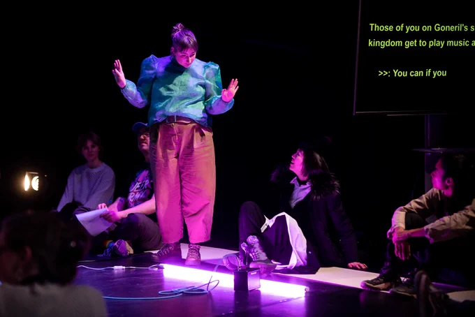 Performer wearing a blue top and brown trousers, surrounded by people sat on the floor and purple glow light at her feet. 