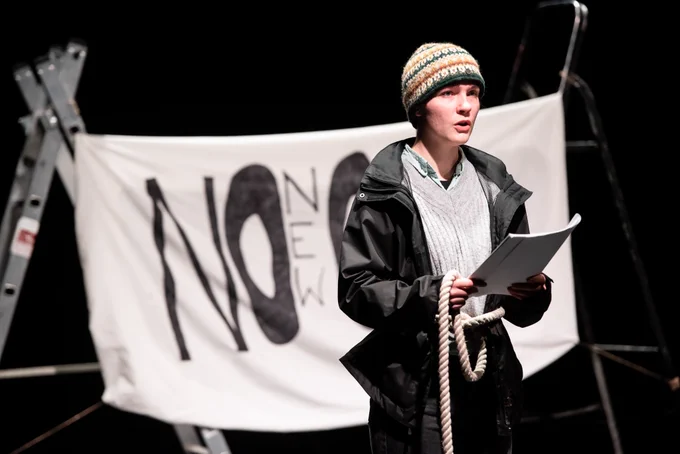 Performer wearing a woolly hat and black jacket reading from a script in front of white sheet behind saying ‘No New Oil’