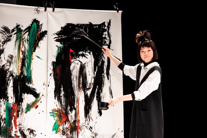 Performer wearing a white shirt and black over dress stood in front of a white canvas with black and red paint on it. 