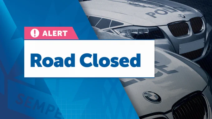 "Alt text: On the left side, a pink and white box with a warning icon and the word "ALERT" is prominently shown. Below this, a large white box contains the text "Road Closed" in bold blue letters. The right side of the image features a blue-tinted, slightly blurred photograph of police cars, indicating the closure is likely due to law enforcement activity or an emergency situation."