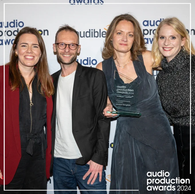 Four white people stand smiling in front of the Audio Production Awards step and repeat. They are wearing smart clothes and celebrating, Charlotte holding the glass trophy for Production Company of the Year.