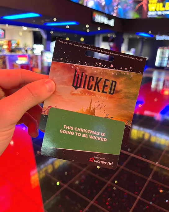 Hand holding a Cineworld gift card designed for the film "Wicked". The card features a green background with the text "This Christmas is going to be Wicked" with a Cineworld logo against a black background at the bottom of the card. The image also features a blurred background of a cinema arcade.