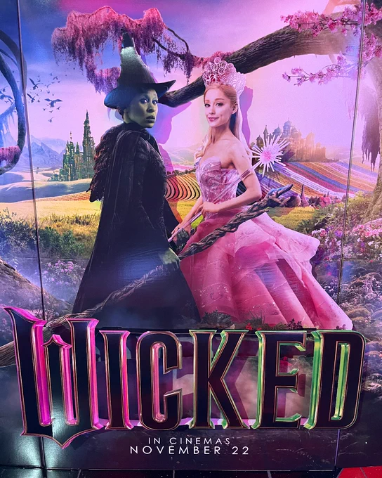 Promotional movie poster for "Wicked," featuring two characters in elaborate costumes standing in a vibrant, enchanting forest setting. The poster announces the movie release date as November 22.