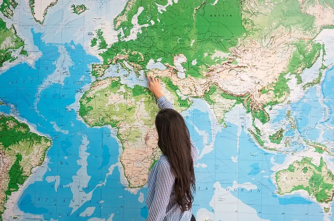 A woman with long brown hair and a blue and white striped shirt pointing to a large map of the world.