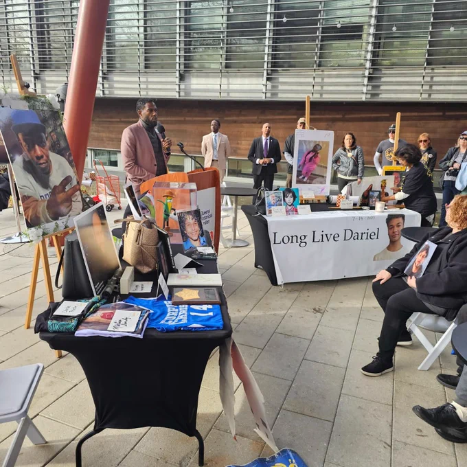 The Public Advocate speaks in an outdoor atrium, next to photos of traffic violence victims and a banner, "Long Live Dariel." Members of the audience hold photos of their loved ones
