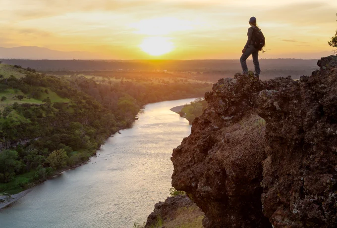 Hiker standing on rock overlooks river with sun setting in the background