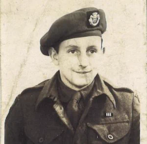 An old, creased black and white portrait photo of a young man in army uniform. He is smiling. He wears a beret with the DCLI insignia. 