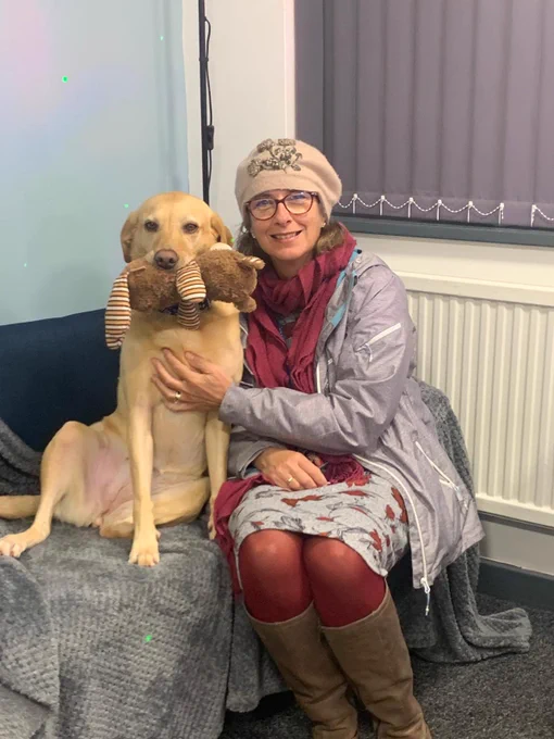 Andrea sat on a seat next to a yellow Labrador called Bella. Bella has a plug toy in her mouth.