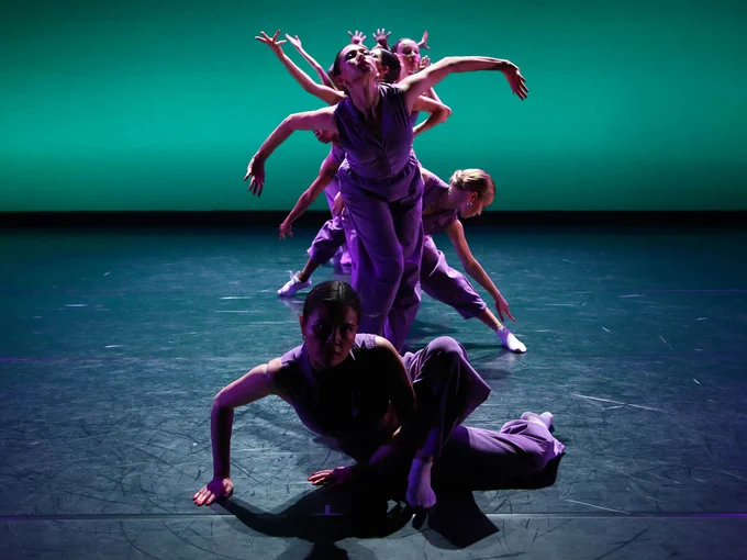 A group of dancers in purple outfits perform on a dimly lit stage with a green background.
