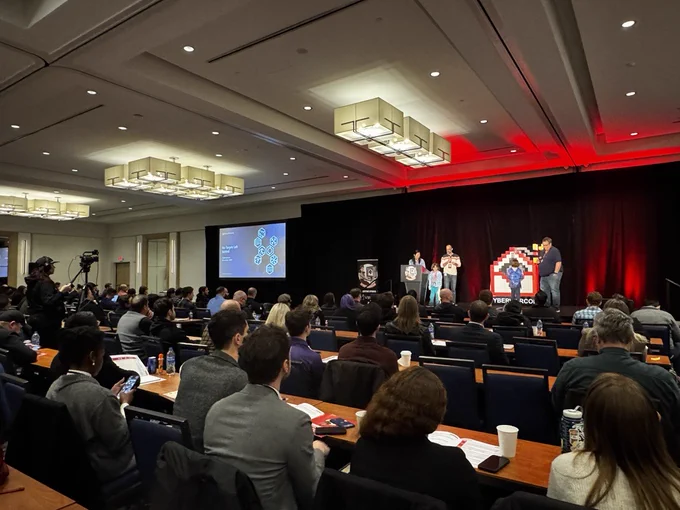 A wide shot showing Judy Ng, Ned Moran, and Mark Parsons standing on the CyberWarCon stage presenting. In front of them is a nearly full seated audience facing them.