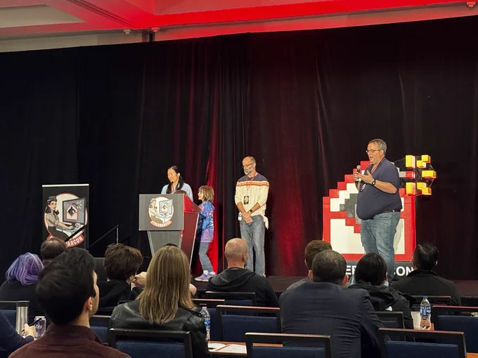 From left to right, Judy Ng, Ned Moran, and Mark Parsons stand on the CyberWarCon stage presenting. Mark Parsons holds a microphone and is speaking.